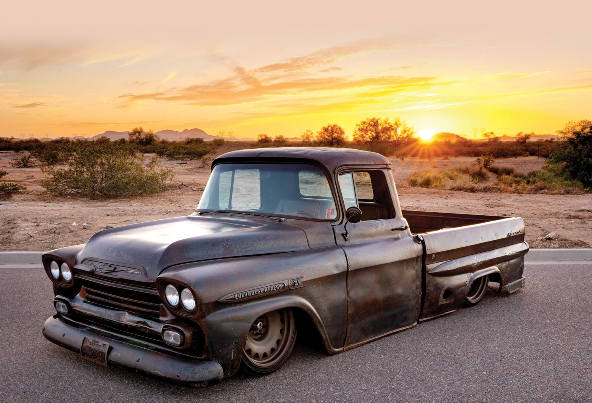 three quarter view of a '59 Apache with a dark patina finish, parked on a desert highway at sunset