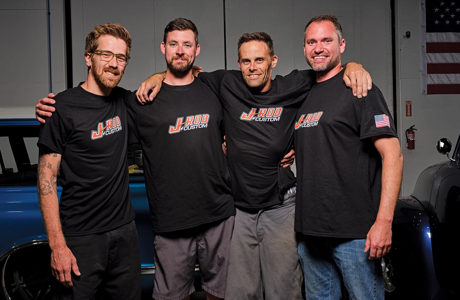 four men stand huddled together in a garage for a photo, each wearing a J-Rod Custom t-shirt