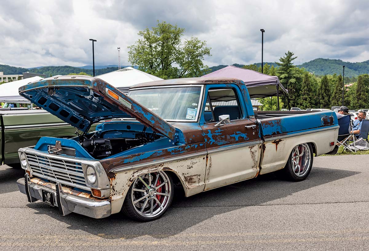 Close-up photograph perspective of a rusty vintage blue/brown/tan Ford F-100 car with its hood open parked next to a green vehicle at the 2023 Grand National F-100 Show in Pigeon Forge, Tennessee with other spectators nearby on a gloomy day