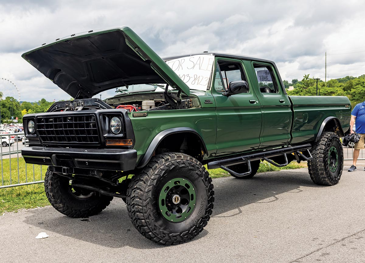 Close-up photograph perspective of a vintage dark green Ford F-350 custom truck with its hood open and a For Sale sign on the front mirror at the 2023 Grand National F-100 Show in Pigeon Forge, Tennessee with one other spectator nearby