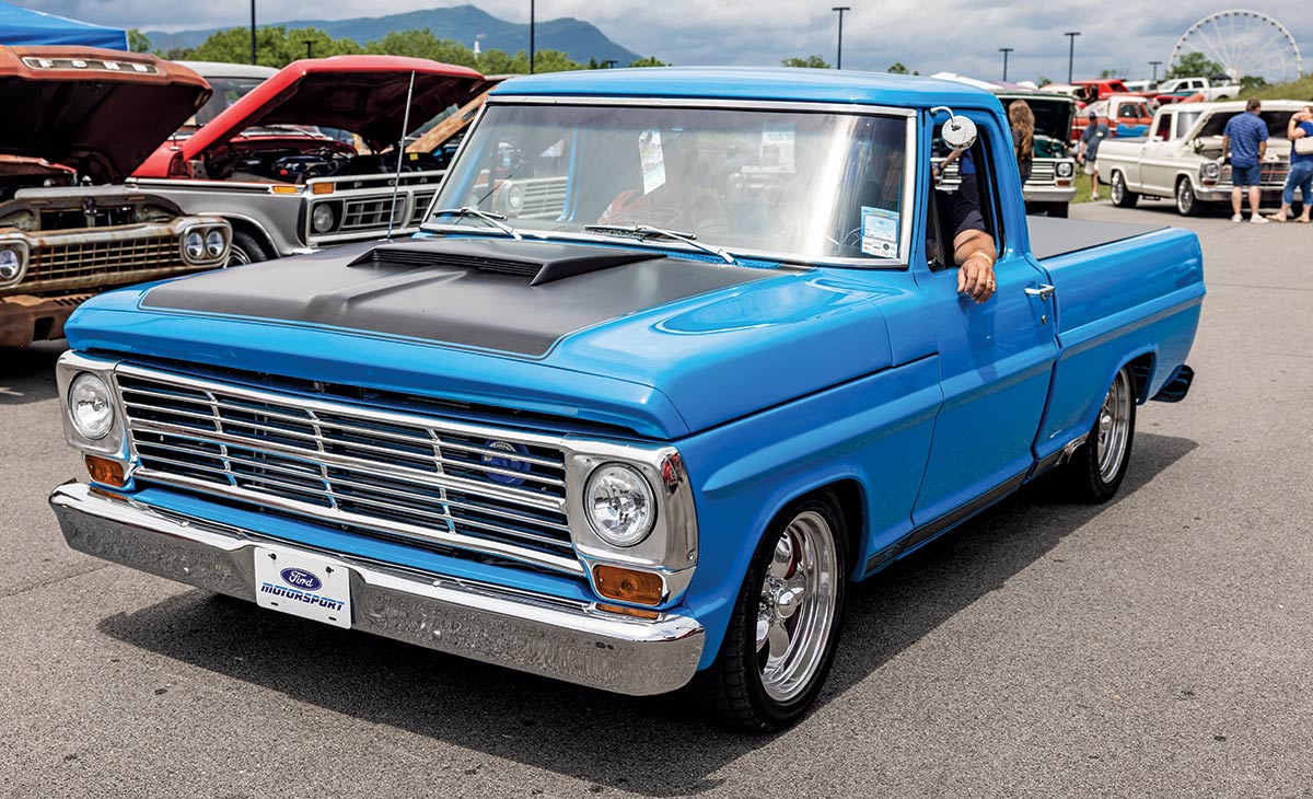 Close-up photograph perspective of a vintage shiny baby blue Ford F-100 car being driven and another passenger inside the car at the 2023 Grand National F-100 Show in Pigeon Forge, Tennessee with other spectators and vehicles nearby