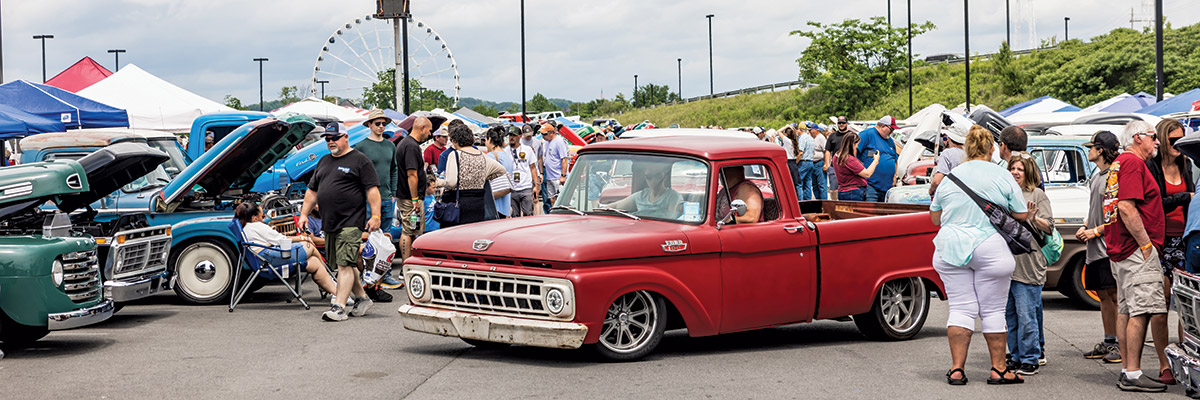 Spectators gather together at the 2023 Grand National F-100 Show in Pigeon Forge, Tennessee as a red vintage Ford F-100 car is seen in the middle of the action with two men inside the vehicle on a gloomy day