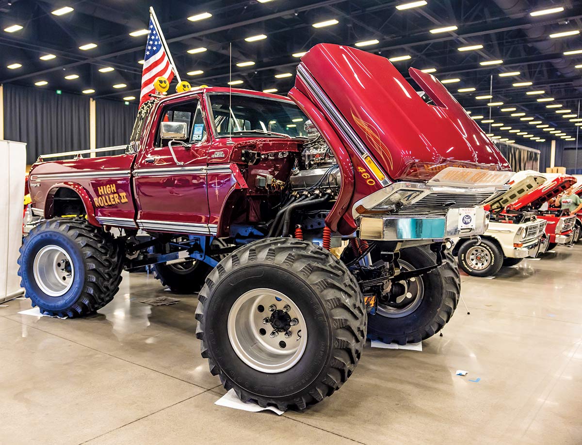 Close-up photograph perspective of a shiny vintage dark red Ford F-350 Ranger car "High Roller II" on display with the hood open parked next to other vehicles inside the 2023 Grand National F-100 Show in Pigeon Forge, Tennessee