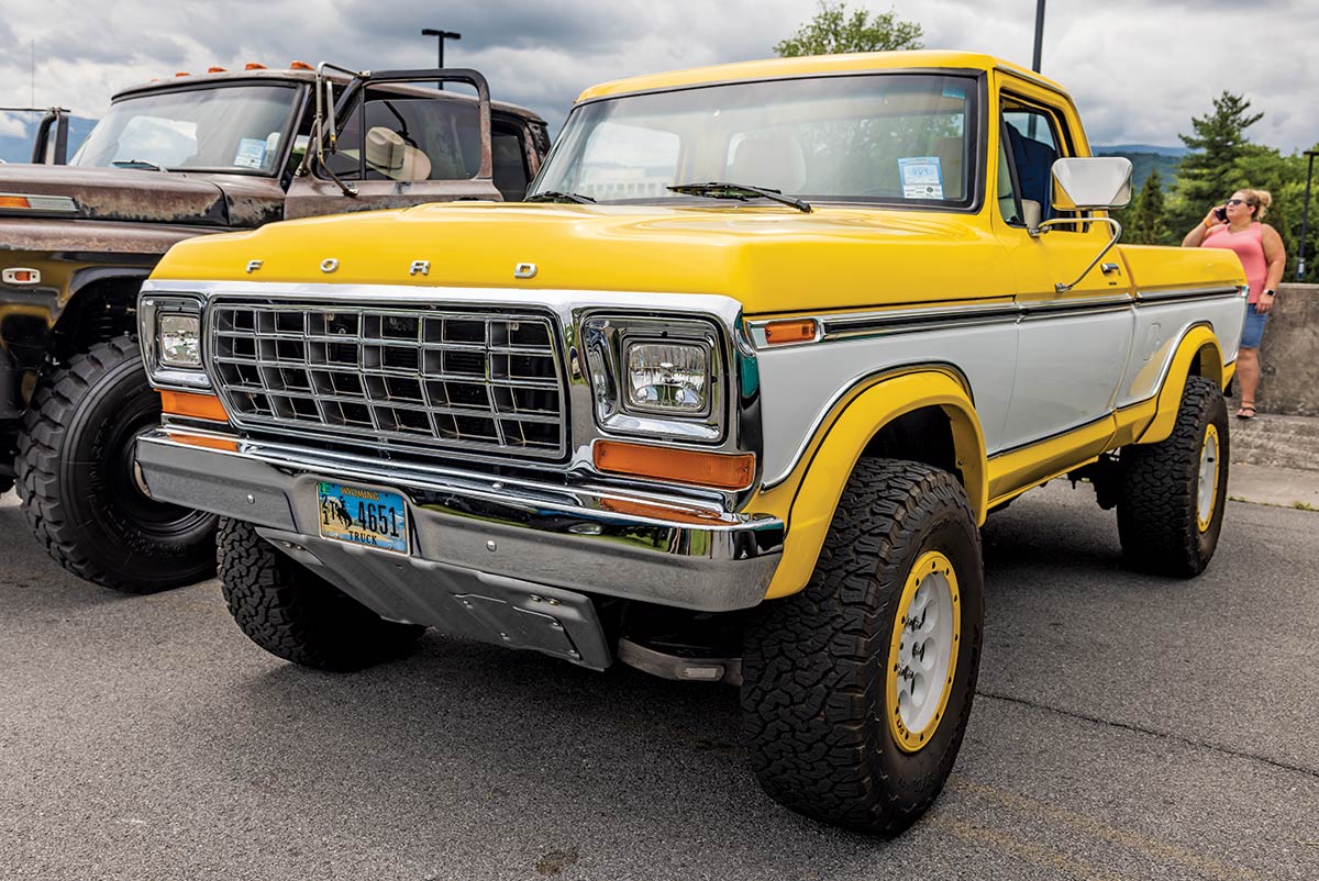 Close-up photograph perspective of a vintage yellow Ford F-600 truck parked next to a rusty old brown and black vintage Ford F-600 truck with one other nearby spectator at the 2023 Grand National F-100 Show in Pigeon Forge, Tennessee on a gloomy day