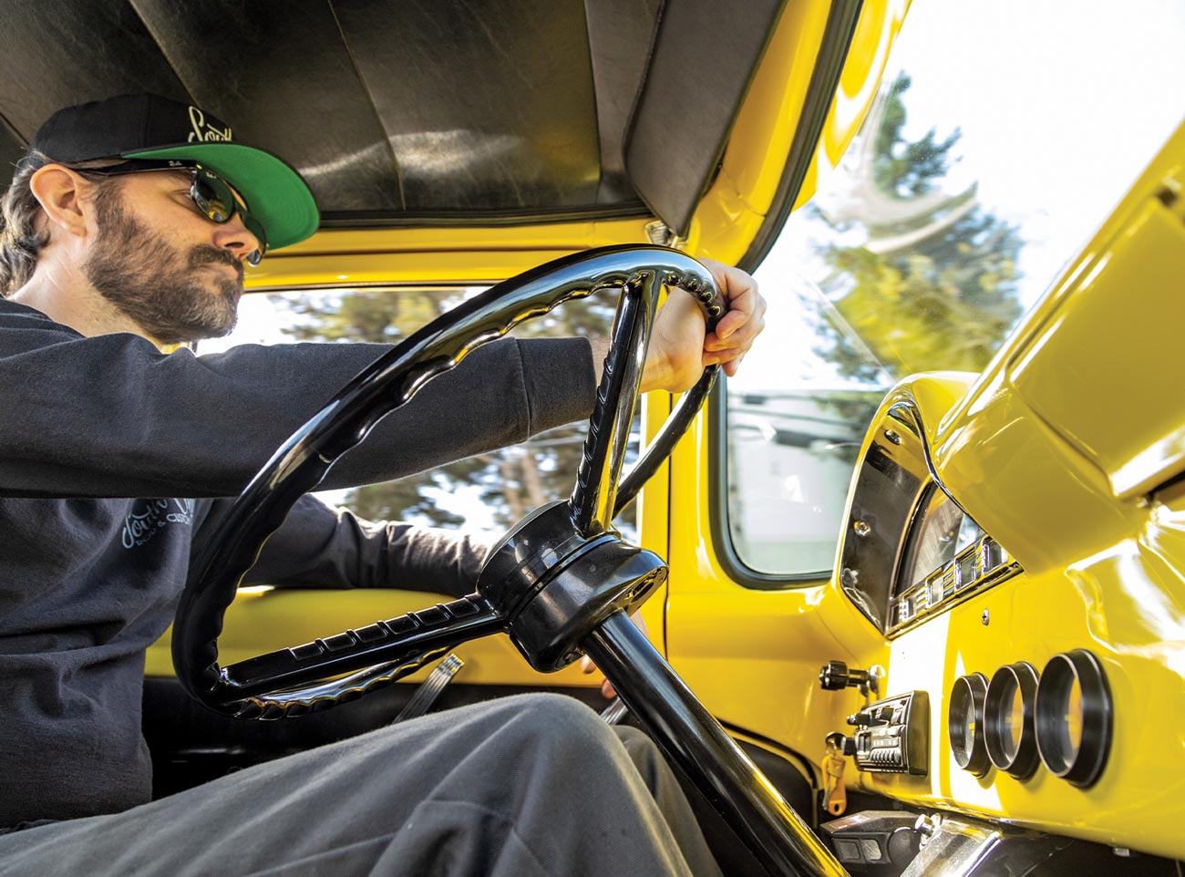 low angle view from the passenger seat of a man, wearing a ball cap, sunglasses and a sweatshirt, at the large classic wheel of the banana yellow ’56 Ford F-100
