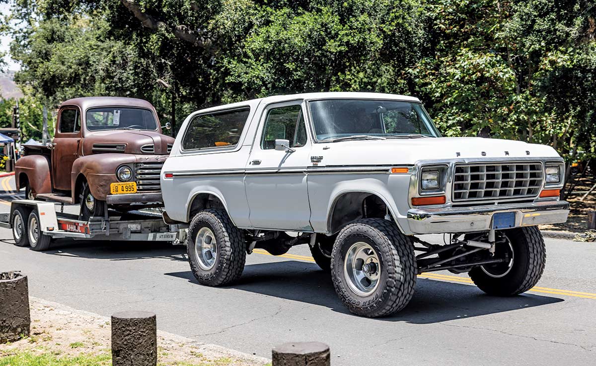 Landscape photograph perspective of a white vintage Ford Bronco hauling a bronze rusty vintage Ford vehicle on a small trailer as it drives on the road at Canyon RV Park in Anaheim, California during the day