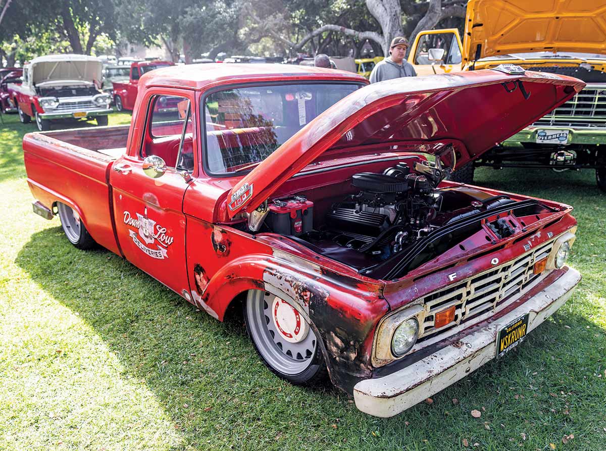 Landscape close-up photograph perspective of a red vintage Ford vehicle "Down Low Deliveries" on display parked on the grass with other spectators nearby roaming around the area and other vehicles parked nearby as well at Canyon RV Park in Anaheim, California