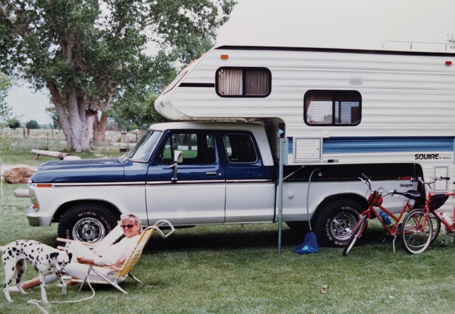 an old dated looking photo of the ’75 Ford SuperCab with a trunk trailer parked on a grassy yard as a woman sits in a lawn chair smiling next to a Dalmatian