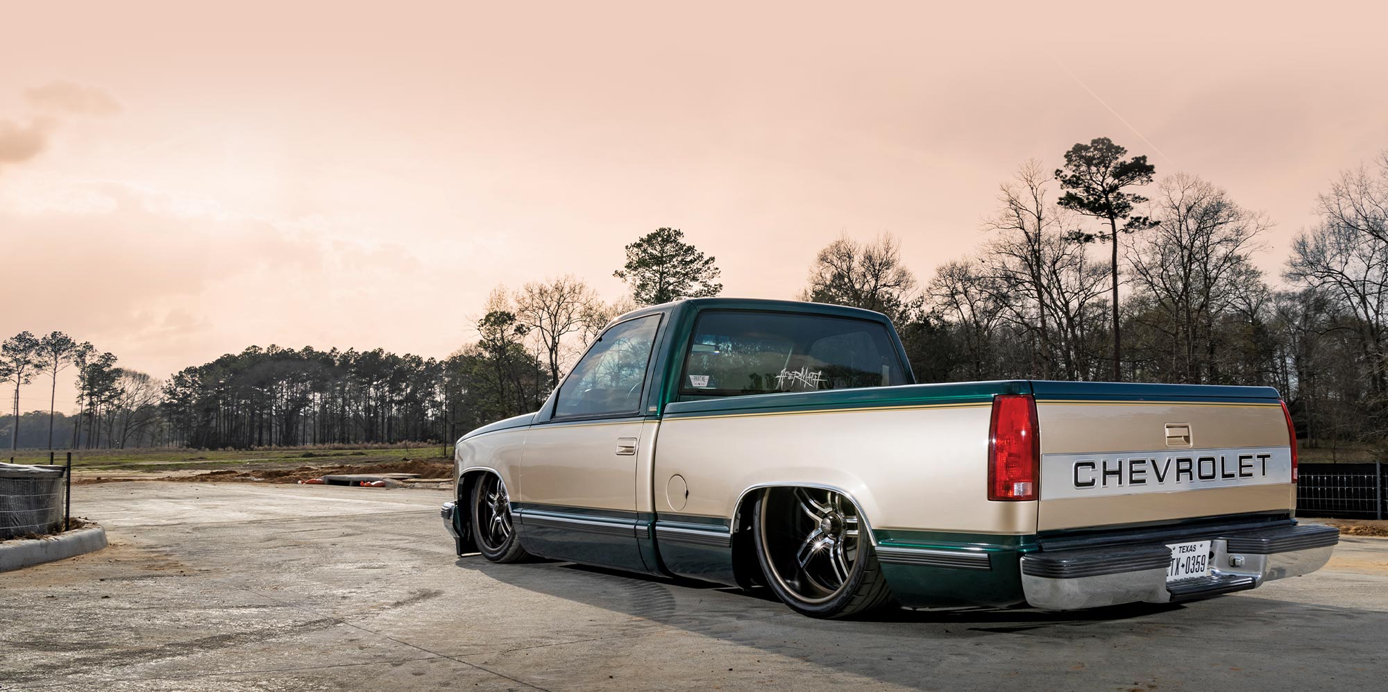 3/4ths rear drivers side view of Jose Galvan’s two-tone dark emerald green and pale gold ’88 Chevy OBS against a background of woods and a sepia sky