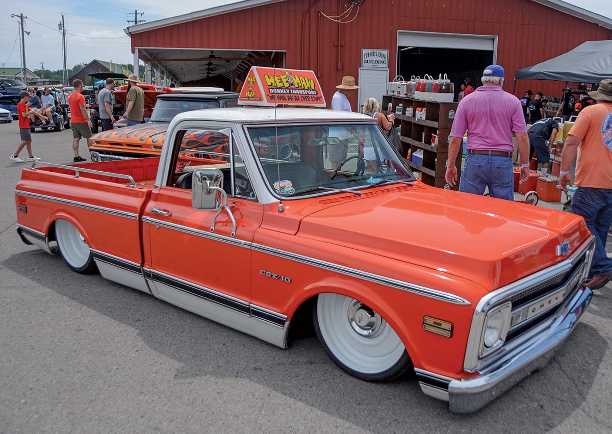 Close-up photograph perspective of a bright shiny orange vintage Chevrolet CST/10 vehicle parked nearby a maroon colored building while there are other people walking around