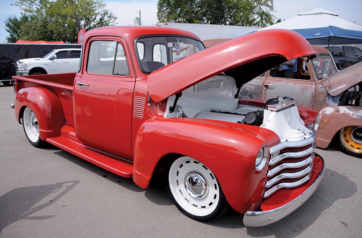 Close-up photograph perspective of a shiny bright red vintage Chevrolet vehicle with its front hood open parked nearby a rusty faded dark brown vintage Chevrolet vehicle with its front hood open as well