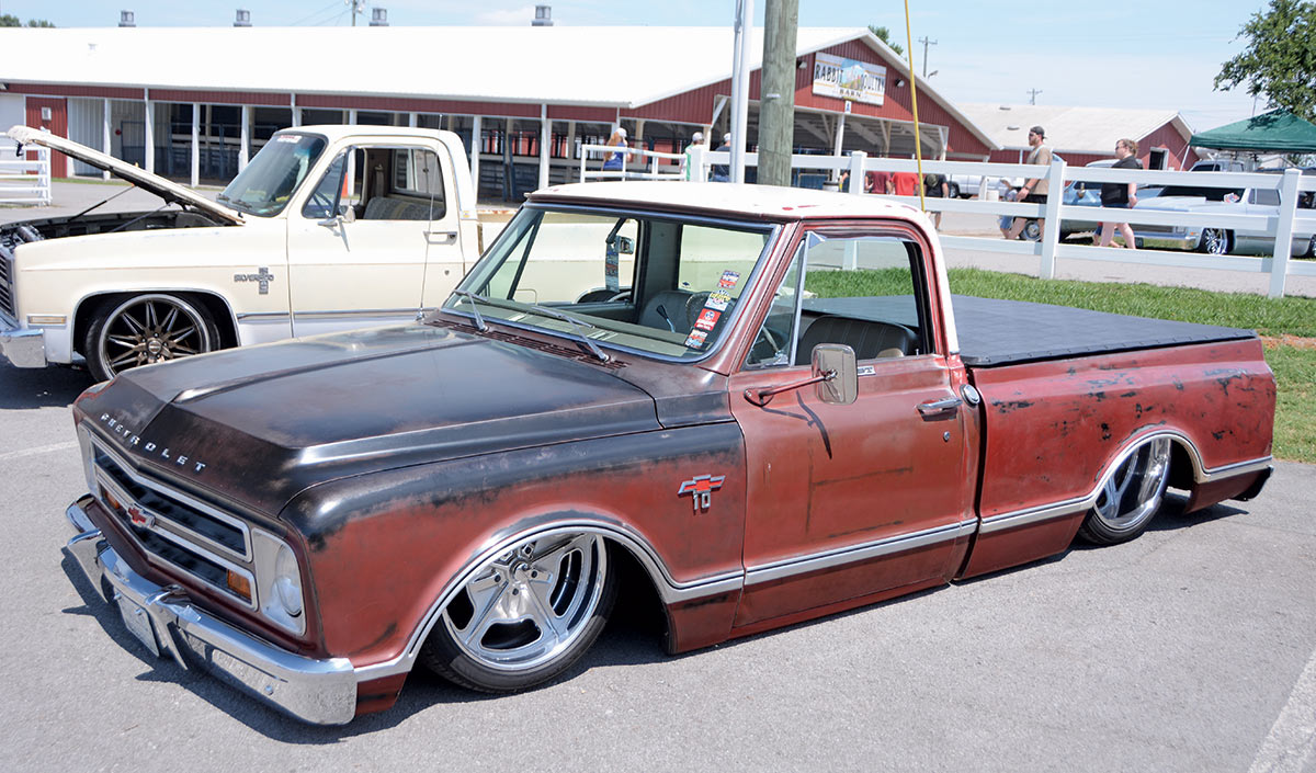 Close-up photograph perspective of a faded dark brown/maroon Chevrolet Custom 10 vintage vehicle parked nearby a tan cream colored vintage Chevy Silverado vehicle with the Silverado's front hood open