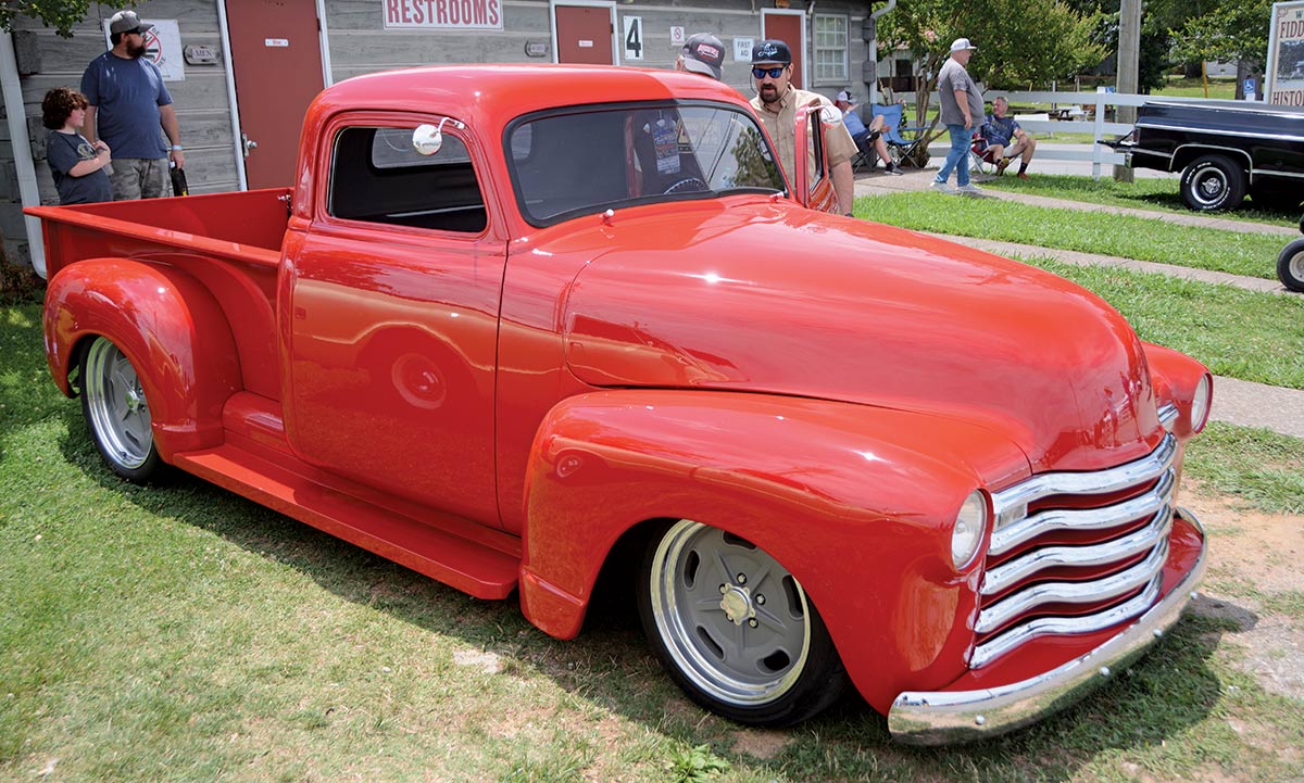 Close-up photograph perspective of a shiny bright red vintage Chevrolet vehicle parked nearby a restrooms building on the grassy lawn as two guys are checking out the driver's side of the car