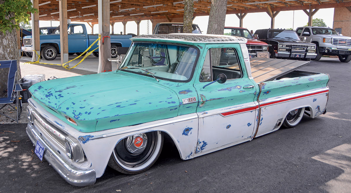 Close-up photograph perspective of a rusty mint green/dark blue vintage Chevrolet C10 vehicle parked nearby a wooden overhead canopy and tree as there is a lawn chair and other cars close by as well