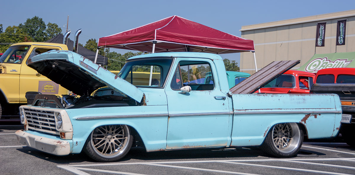 Close-up landscape photograph side perspective of a rusty sky blue-colored Ford vintage vehicle with its hood open parked in a parking lot area nearby other random assorted cars plus a red tent next to it