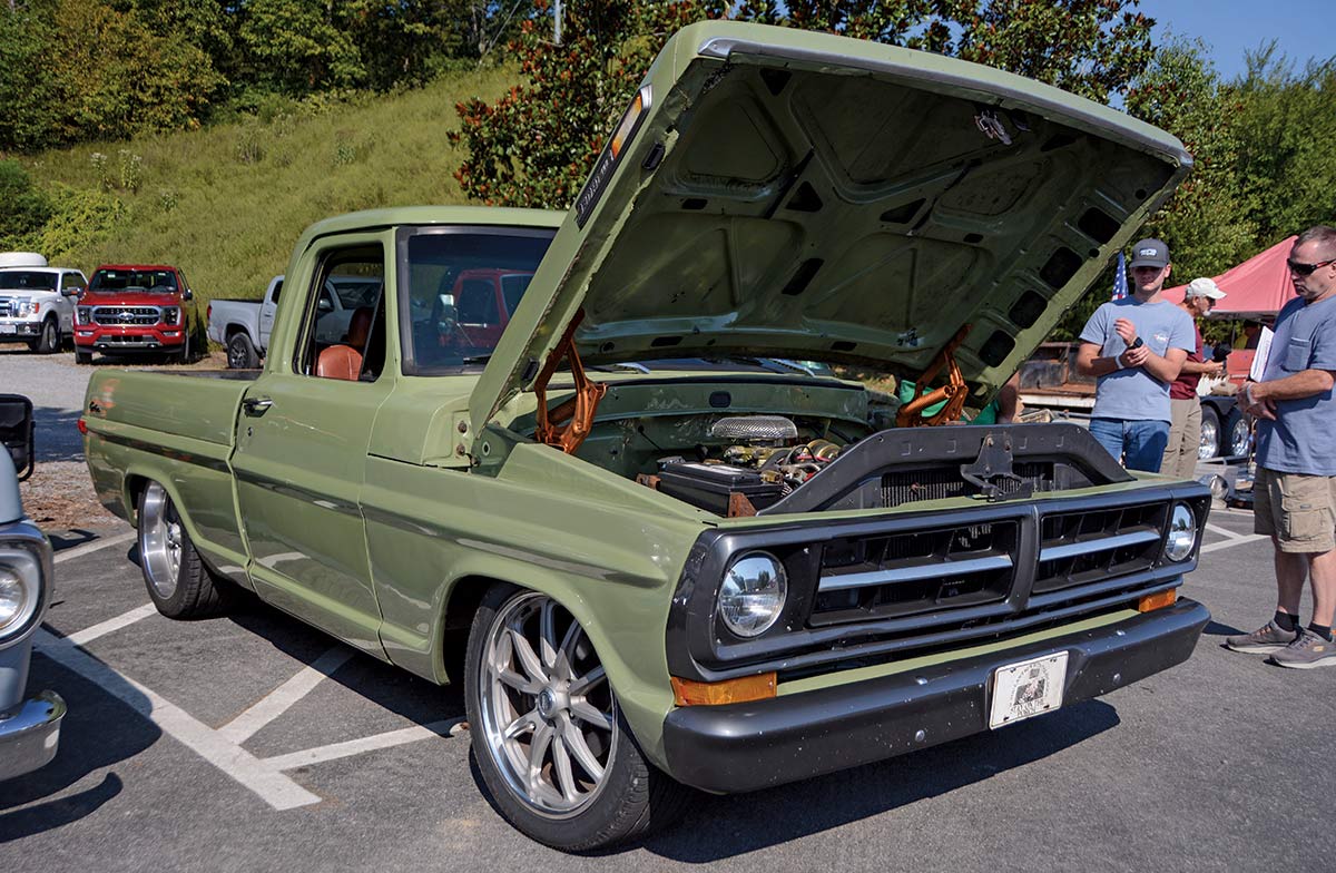 Close-up landscape photograph angle perspective of forest green-colored Ford vintage vehicle with its hood open parked in a parking lot area while two men examine the front hood area from a glance