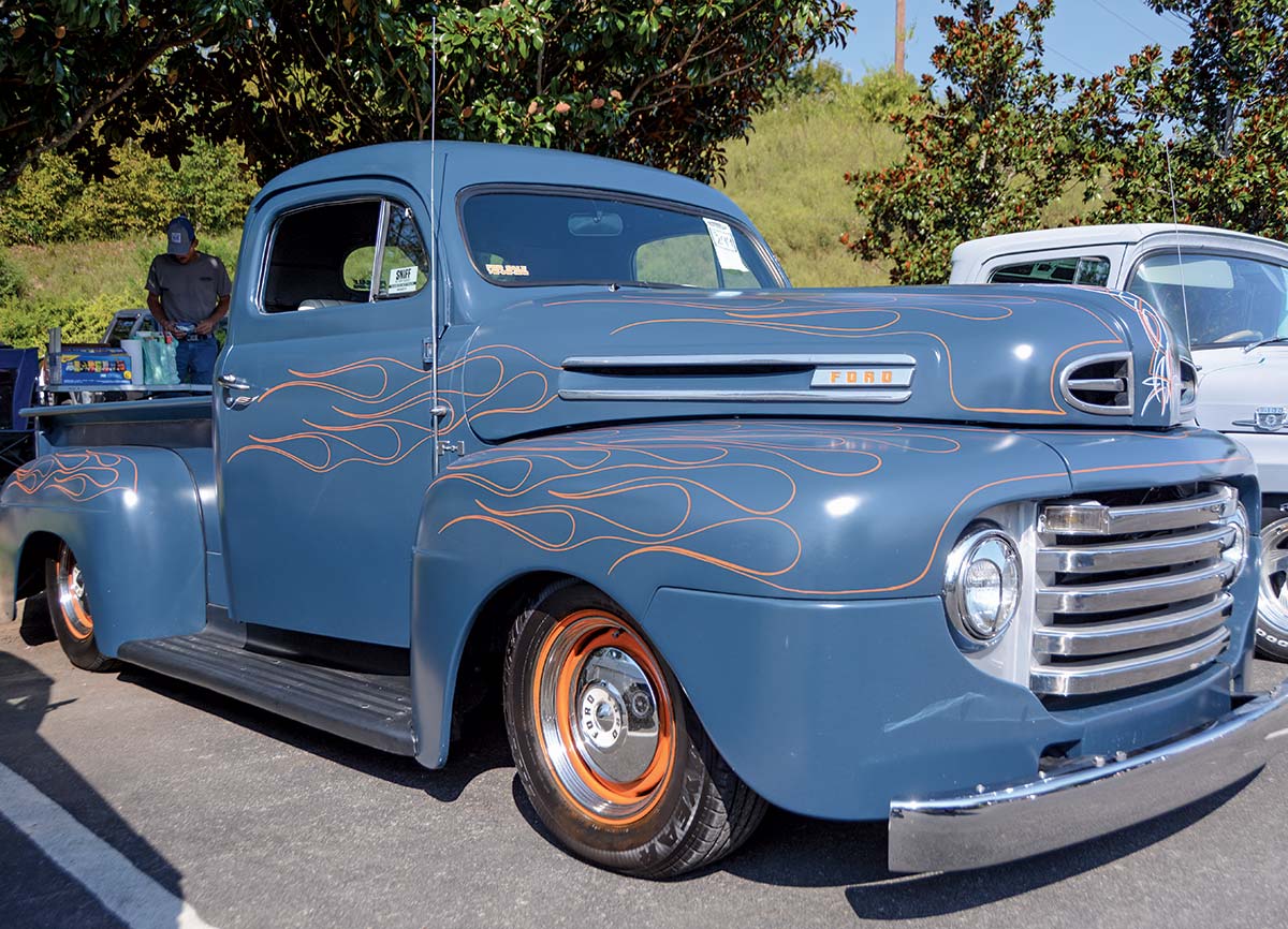 Close-up landscape photograph angle perspective of a blue-colored Ford vintage vehicle parked in a parking lot area next to another random assorted car as there is a man standing in the way far distant nearby a tree