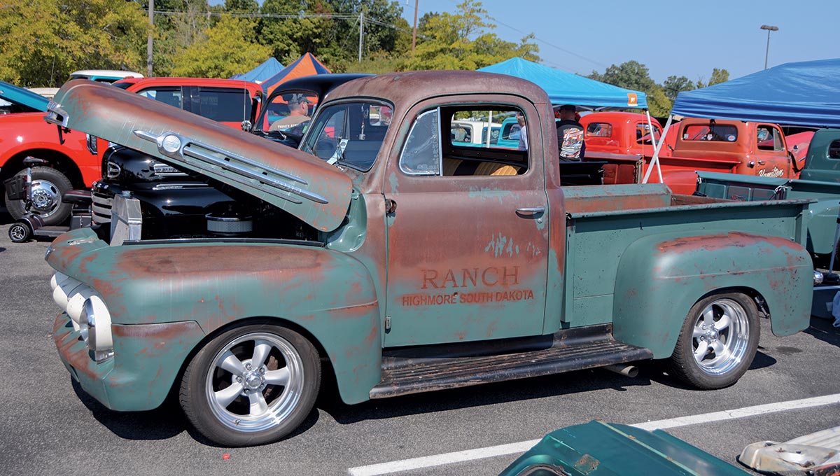 Close-up landscape photograph side perspective of a rusty dark green-colored Ford vintage vehicle with its hood open parked in a parking lot area nearby other random assorted vehicles and tents all around