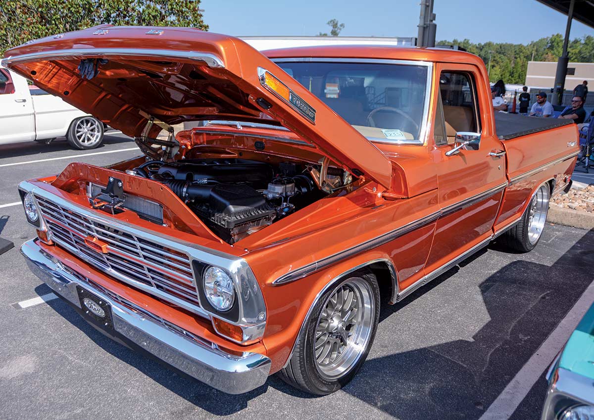 Close-up landscape photograph angle perspective of a shiny/glossy orange-colored Ford vintage vehicle with its hood open parked in a parking lot area next to another random assorted vehicle while there are other people in the far distant background