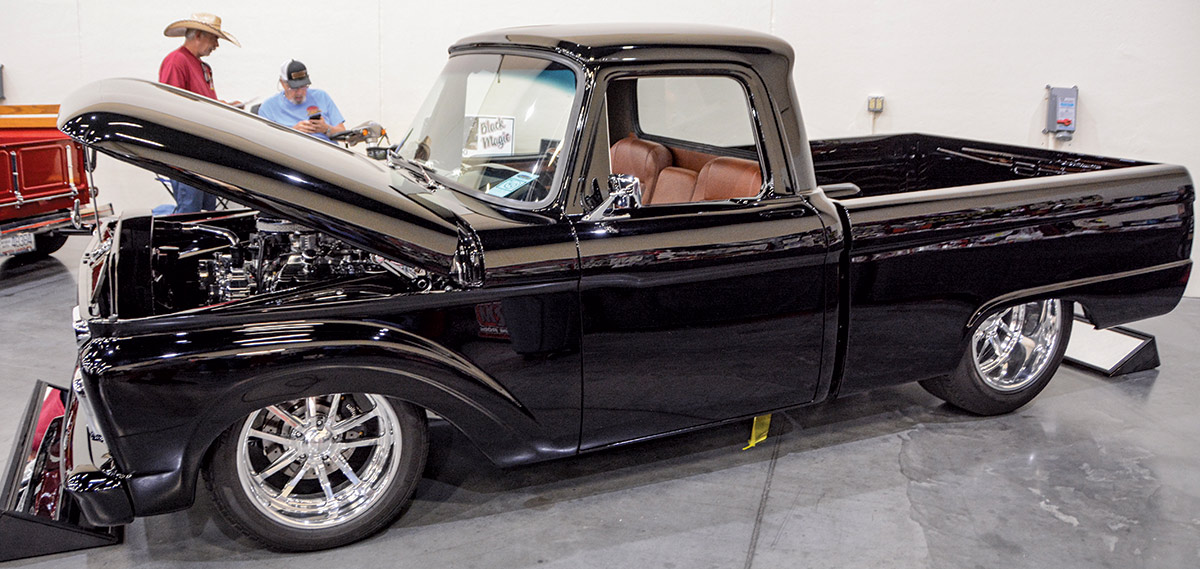 Close-up landscape photograph angle perspective of a shiny/glossy black-colored Ford vintage vehicle with its hood open and there are other people in the way distant background talking to each other