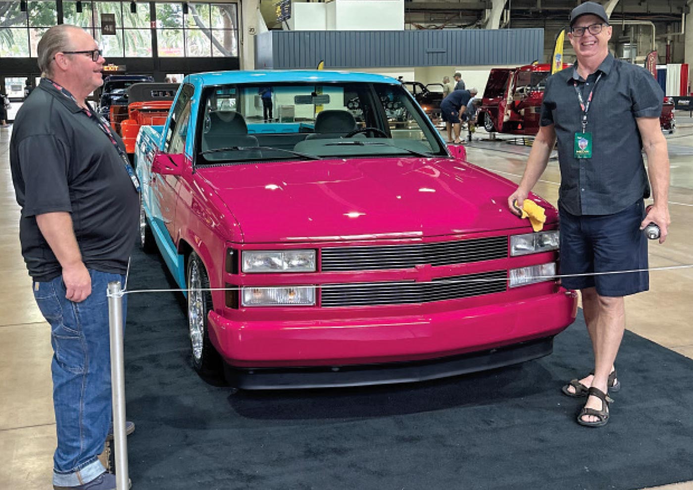 two men stand on either side of a magenta and baby blue classic truck