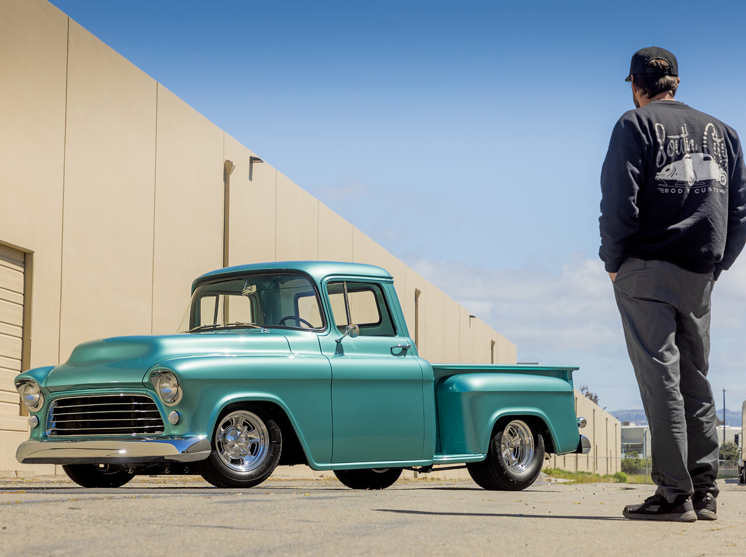 back view of a man admiring the sea green ’56 Chevy truck parked in an empty lot beside a large factory