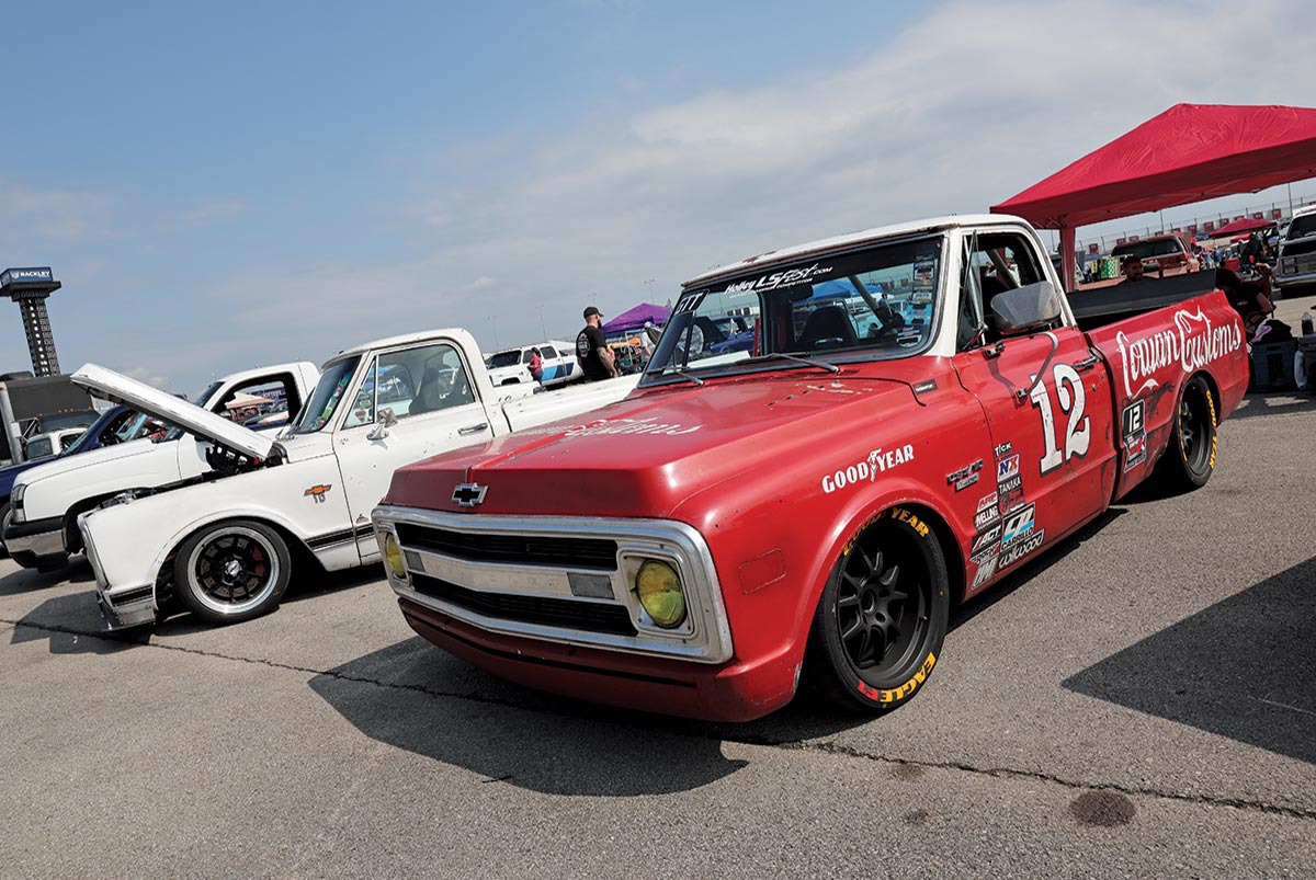 Landscape outdoor photograph of a red/white vintage custom-made Chevrolet C10 truck model parked in a parking lot area nearby other random assorted vehicles and a red tent behind the red/white vintage Chevrolet C10 truck model in the background