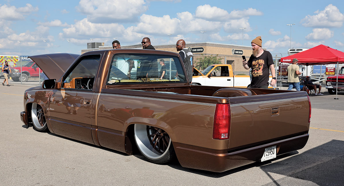 Landscape outdoor photograph of a dark brown/bronze vintage Chevrolet C10 truck model parked in a parking lot area while several men are standing seen checking out the model in the background