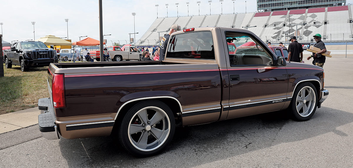 Landscape outdoor photograph of a dark brown/bronze vintage Chevrolet C10 truck model parked in a parking lot area while a man is seen glancing at the model in the background plus another man in the foreground is checking out the driver's side