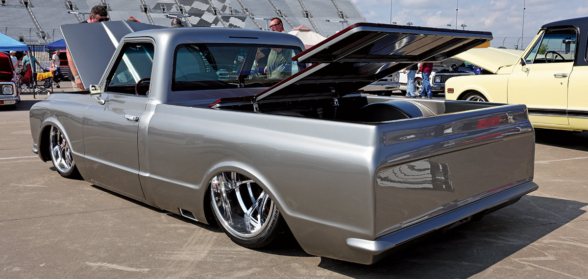 Landscape outdoor photograph of a light grey vintage Chevrolet C10 truck model parked in a parking lot area with its front hood and back trunk slightly open and exposed as a man in the foreground is seen checking out the front hood