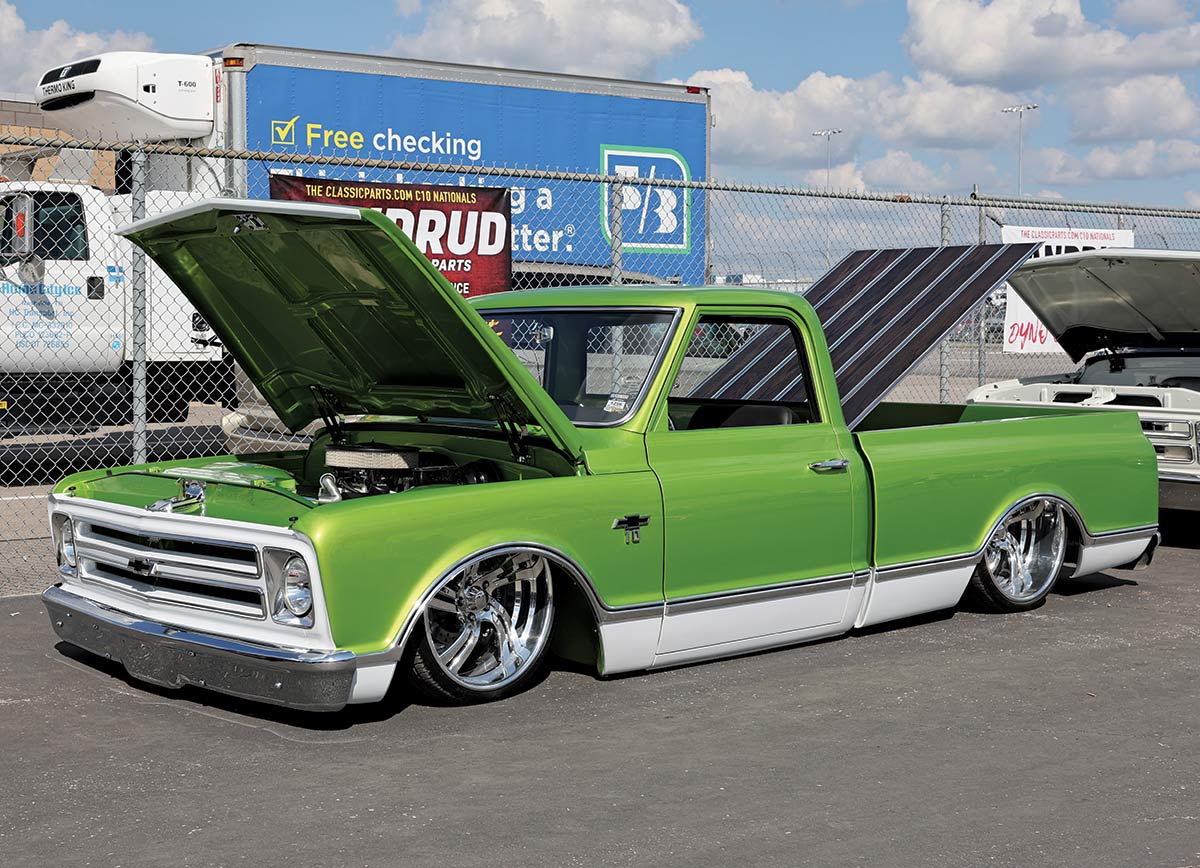 Landscape outdoor photograph of a lime green vintage Chevrolet C10 truck model parked nearby a fence with its front hood and back trunk slightly open and exposed as there are two other random assorted vehicles nearby