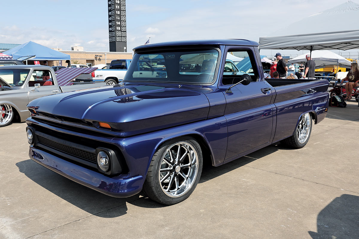 Landscape outdoor photograph of a dark navy blue vintage Chevrolet C10 truck model parked in a parking lot area nearby other random assorted vehicles and other random assorted tents while there are people standing or sitting in the background