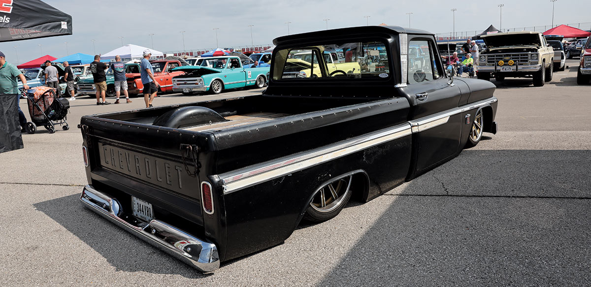Landscape outdoor photograph of a black vintage Chevy C10 truck model parked in a parking lot area with other random assorted vehicles and people roaming around in the background