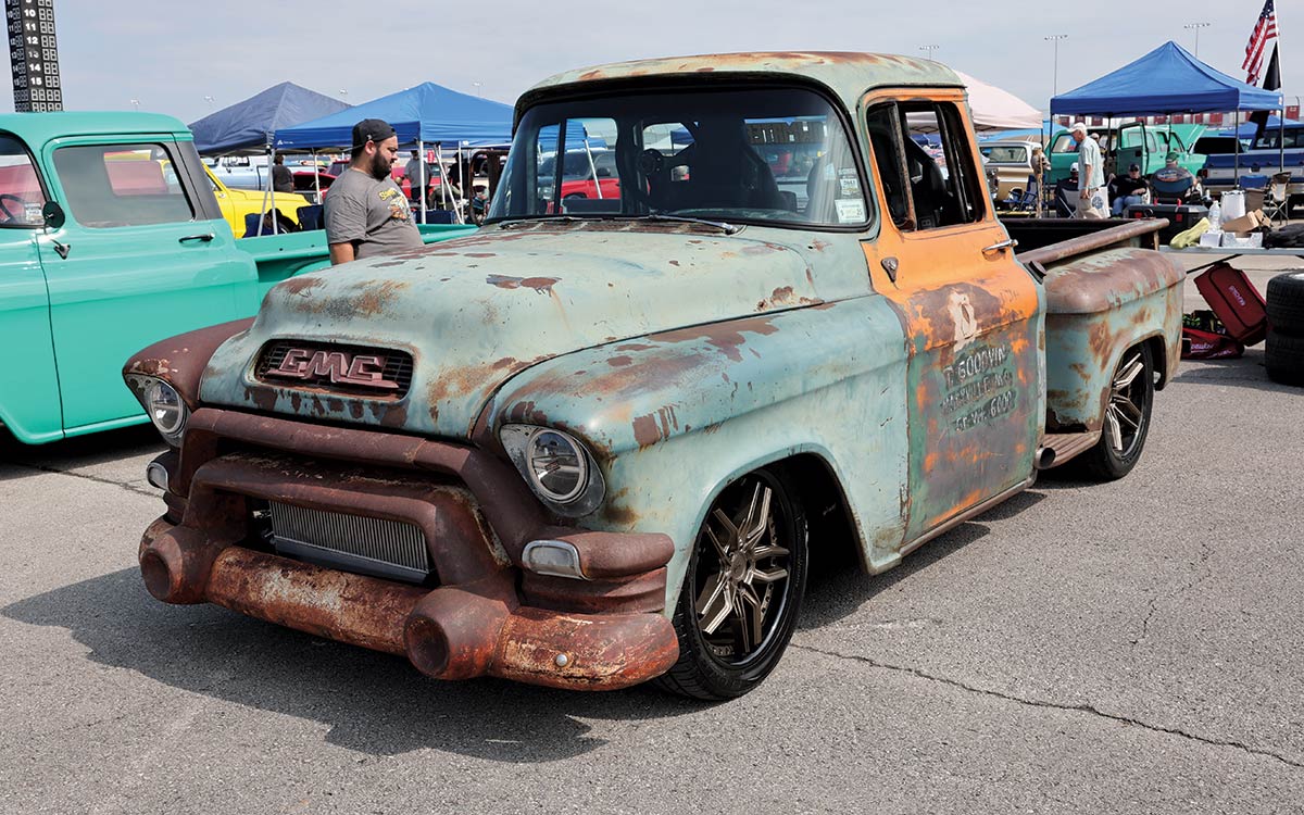 Landscape outdoor photograph of a faded light green/dark brown vintage rusty GMC truck model parked in a parking lot area with a man standing nearby and other random assorted tents in the background