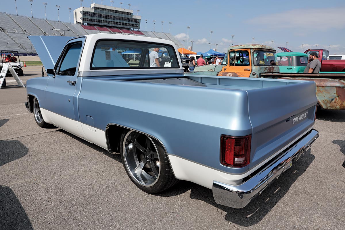 Landscape outdoor photograph of a light sky blue/white vintage Chevrolet C10 truck model parked nearby a racetrack area with other random assorted vehicles parked next to it and other people walking around in the background