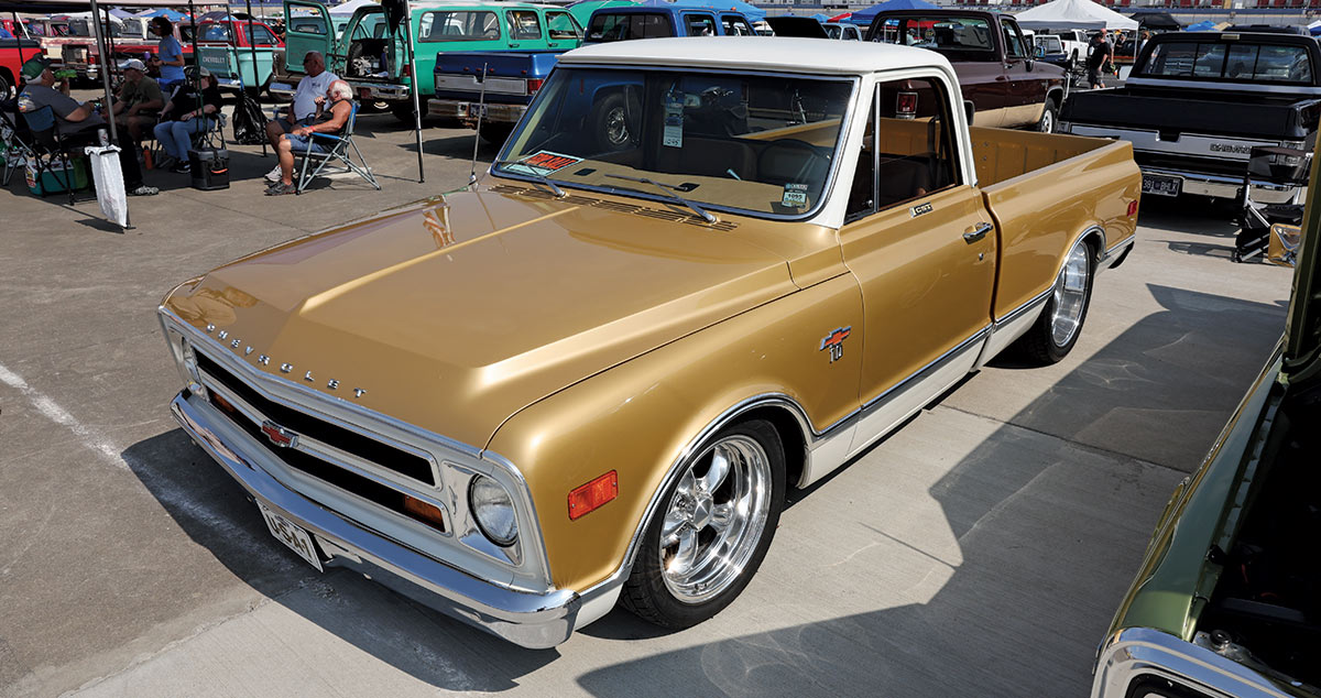 Landscape outdoor photograph of a dark gold vintage Chevrolet C10 truck model parked in a parking lot area with other random assorted vehicles parked nearby and other people sitting down a few yards away in the background