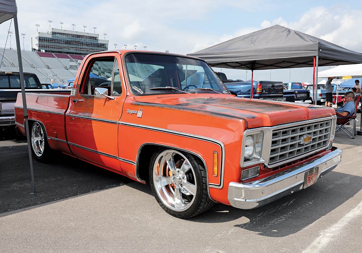 Landscape outdoor photograph of a orange vintage Chevrolet C10 truck model parked in a parking lot area next to a dark grey tent and other random assorted vehicles nearby and a few people in the background