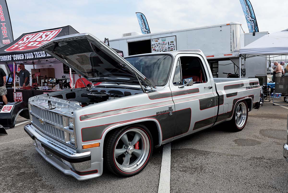 Landscape outdoor photograph of a light grey/black/red vintage Chevrolet C10 truck custom deluxe model parked in a parking lot area with its front hood slightly open and exposed nearby a random logo branded tent
