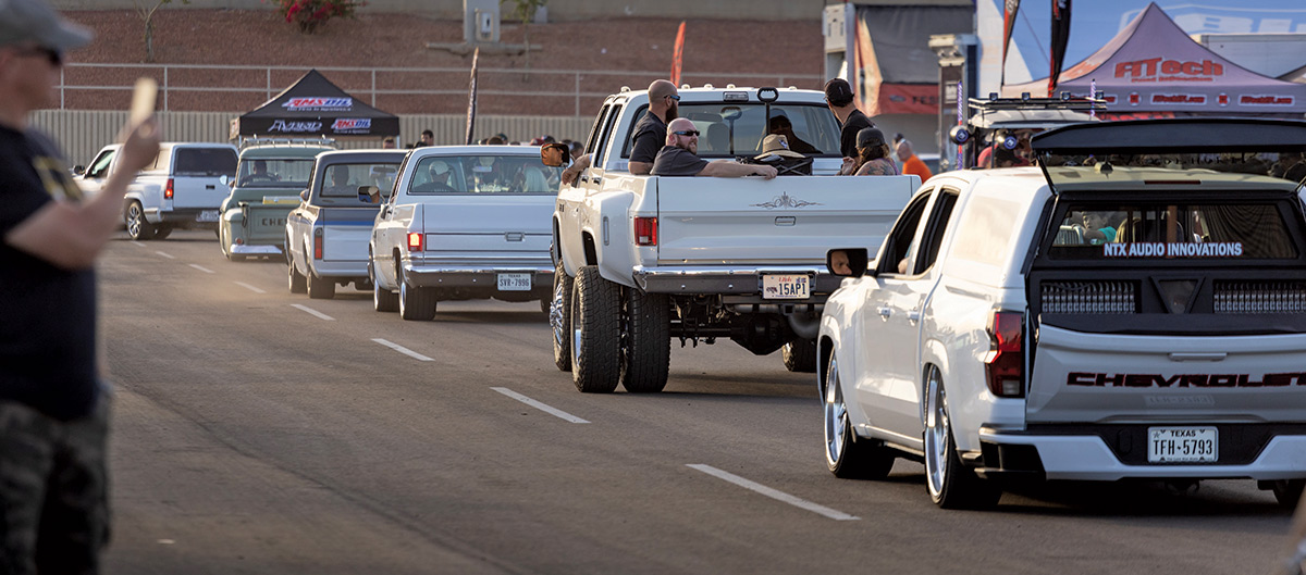 Close-up landscape photograph view of different colored Chevrolet vintage truck vehicles all driving away as there is one vehicle that has several people in it riding in the back trunk which the car's position is located second to last