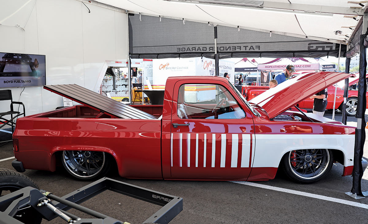 Close-up landscape photograph view of a red/white matte colored Chevrolet vintage truck vehicle with its front hood slightly open and back trunk slightly open as it parked in a parking lot space underneath a Fat Fender Garage logo branded tent