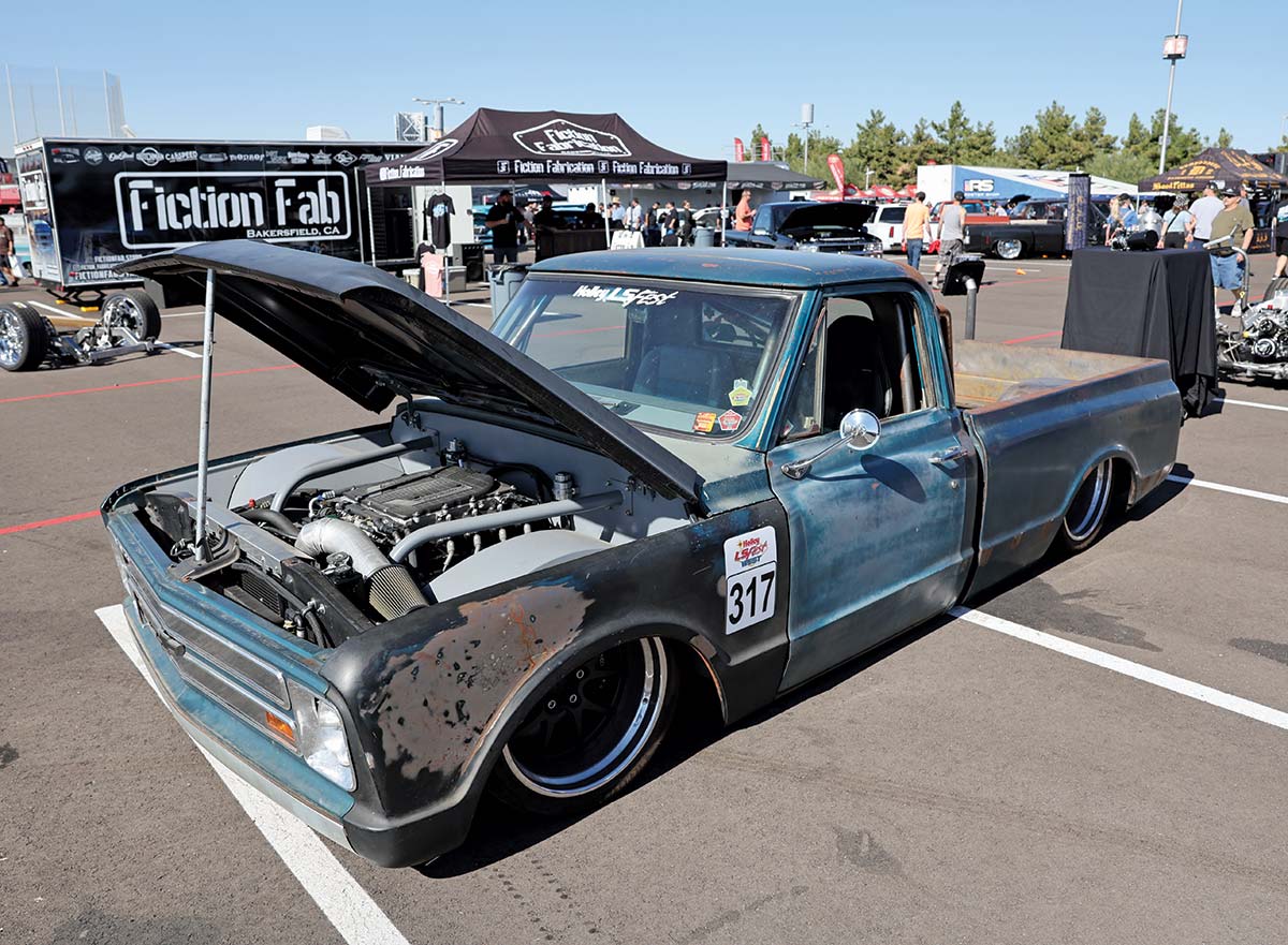 Close-up landscape photograph view of a rusty faded turquoise/black Chevrolet vintage truck vehicle with its front hood slightly open parked in a parking lot space nearby other tents