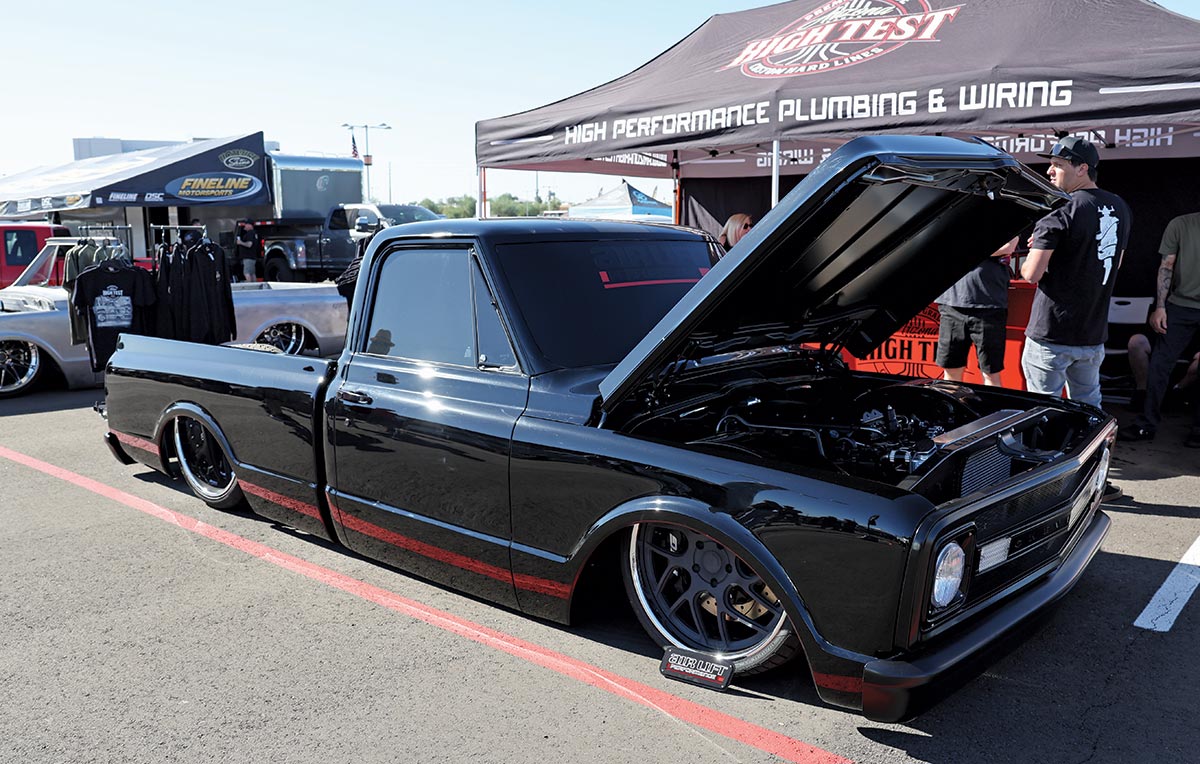 Close-up landscape photograph view of a black matte colored Chevrolet vintage truck vehicle that has its front hood slightly open parked in a parking lot space nearby a High Test Performance Plumbing & Wiring logo branded tent