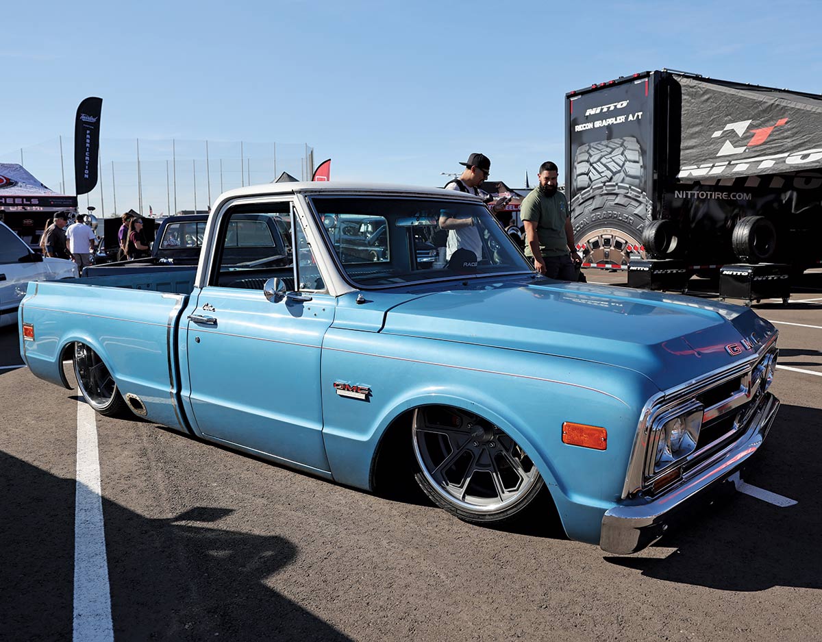 Close-up landscape photograph view of a blue colored GMC vintage truck vehicle parked in a parking lot space as there is other people nearby and a Nitto Tire logo branded trailer and tent
