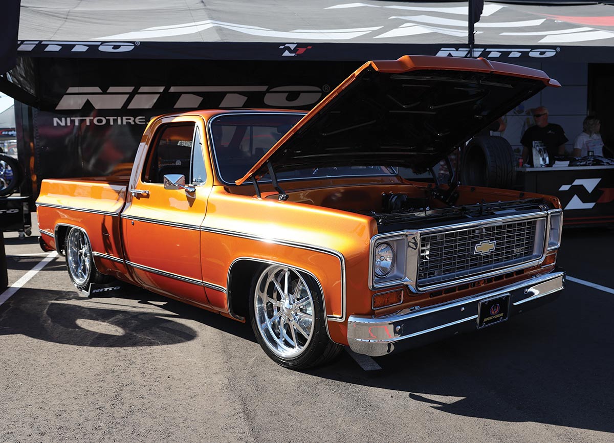 Close-up landscape photograph view of a bright orange colored matte Chevrolet vintage truck vehicle that has its front hood slightly open as the car is parked nearby a Nitto Tire logo branded tent