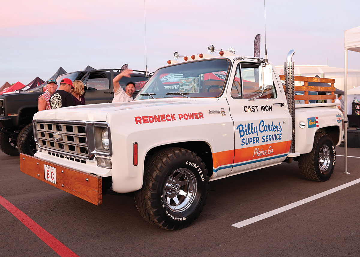 Close-up landscape angle photograph view of a white/orange/wood brown custom-themed Chevrolet C10 vintage truck vehicle titled Billy Carter's Super Service parked in a parking lot space area as a man is seeing waving and posing next to the passenger side of the vehicle