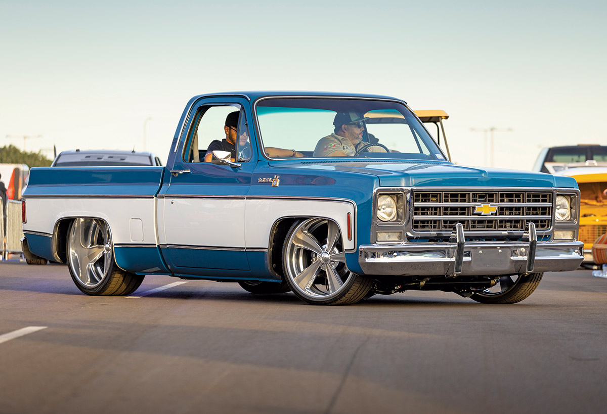 Close-up landscape angle photograph view of a blue/white Chevrolet C10 vintage truck vehicle parked with a man in sunglasses and a hat in the driver's seat while opposite him is a passenger