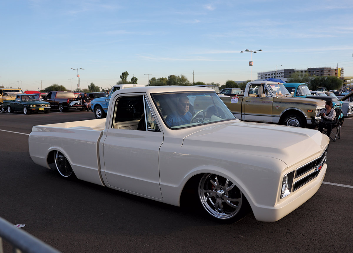 Close-up landscape angle photograph view of a dark cream Chevrolet C10 vintage truck vehicle parked with a man in sunglasses in the driver's seat while opposite him is a passenger