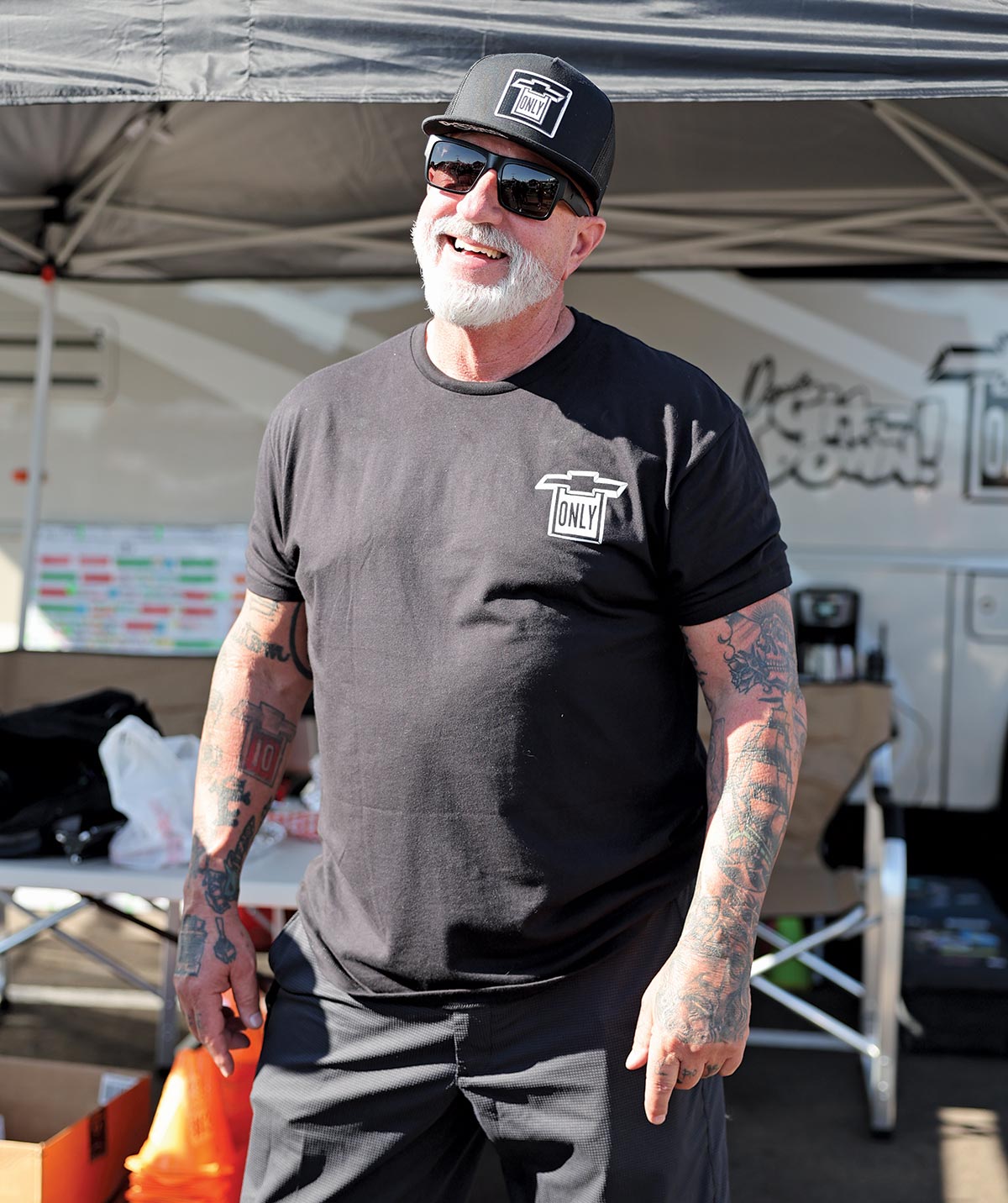 Close-up portrait photograph view of a man smiling as he is wearing a black Chevy only branded logo graphic t-shirt and a black hat with the same Chevy only branded logo on it