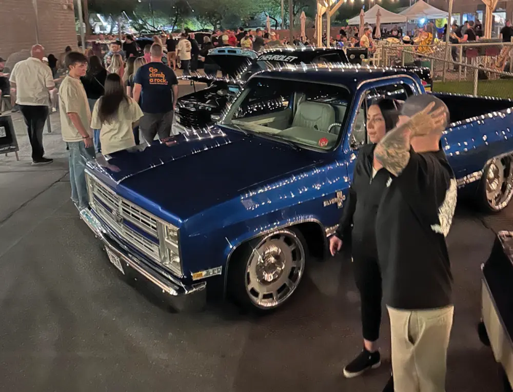 a man and a woman stand beside a dark metallic blue classic Chevy Silverado, taking in the busy late evening scene at the 2024 Reunion Truck Show