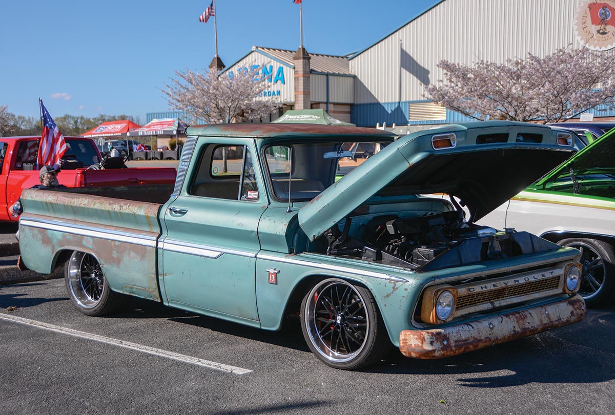 Landscape outdoor close-up photograph angle view of a vintage rusty faded dark turquoise Chevrolet C10 truck vehicle parked in a parking lot area nearby other random assorted cars during the day as there's a big building and other various tents in the distant background